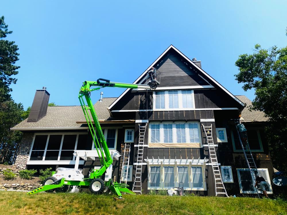 Workers using a lift to paint a tall, modern house with green accents on a sunny day.