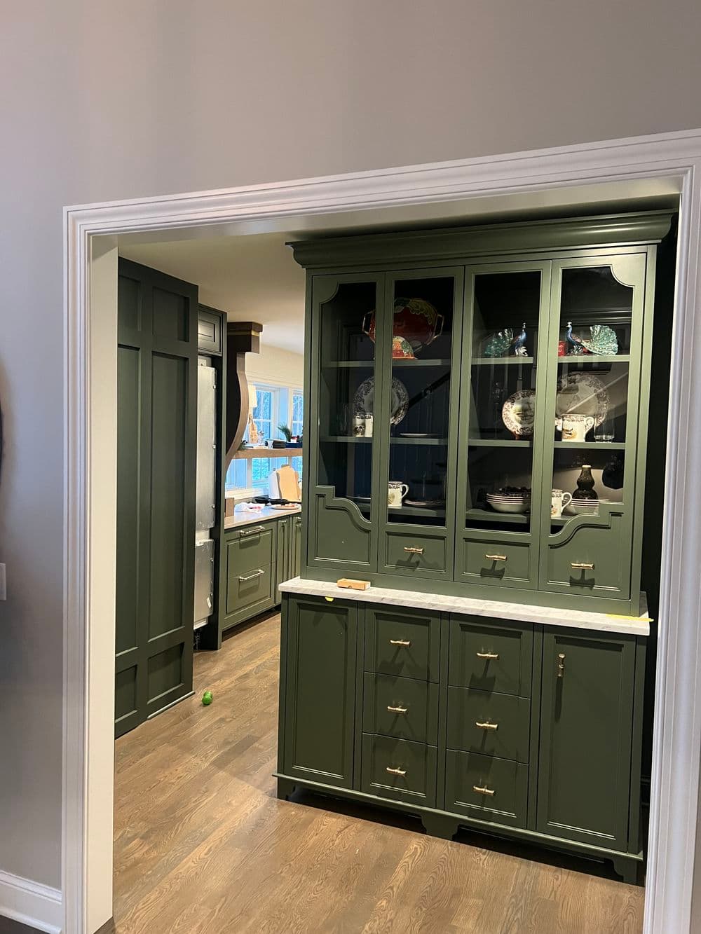 Elegant green kitchen cabinetry with glass doors and marble countertop, showcasing dishware.