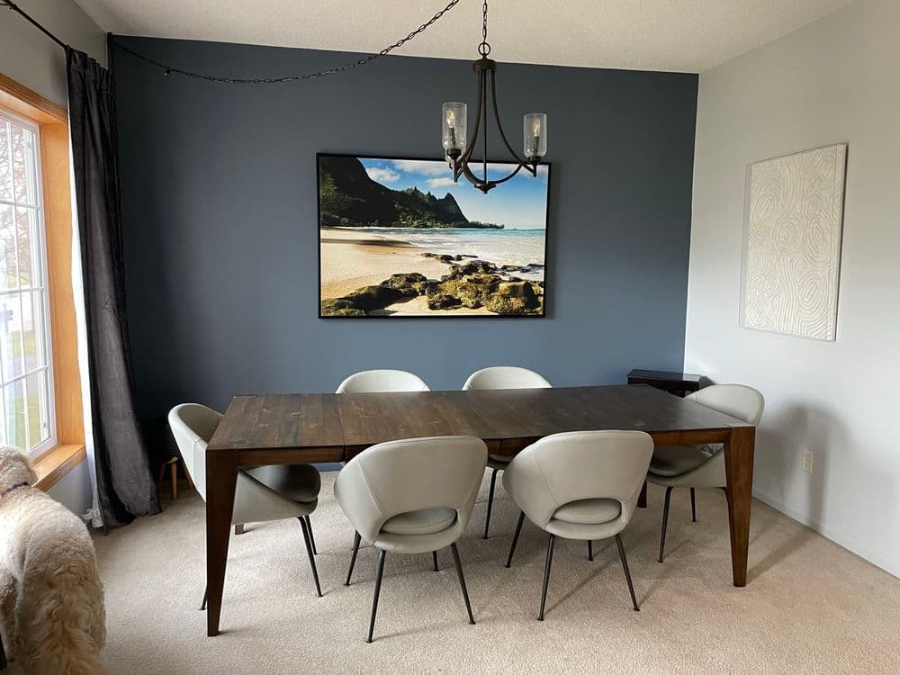 Modern dining room with a wooden table, six gray upholstered chairs, and a coastal artwork.