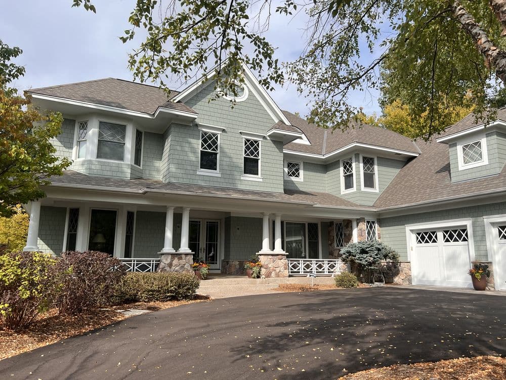 Elegant green house with porch, landscaped yard, and driveway surrounded by trees.