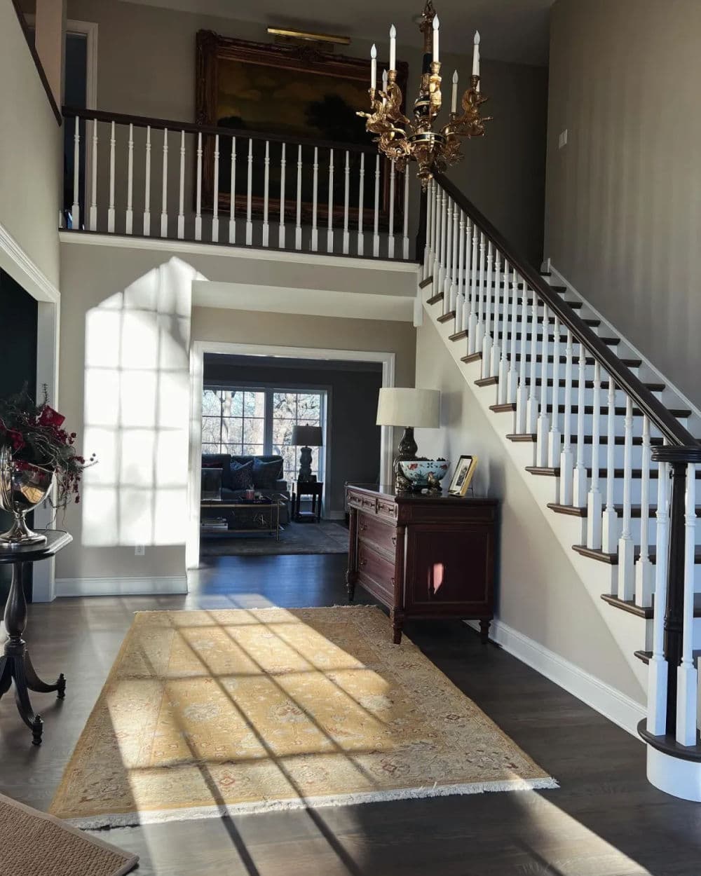 Elegant foyer with staircase, chandelier, and natural light casting shadows on a rug.